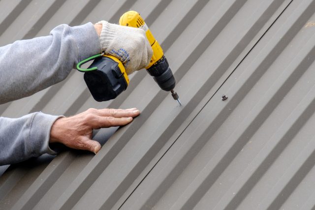 Male hands in gloves with a screwdriver screw the roofing sheet to the roof.